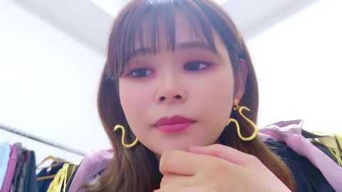 Media: Video of an Asian woman with long brown hair, wearing large golden hoop earrings, pink lipstick, and a purple jacket, sitting indoors with a blurred background.