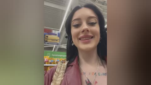 Media: Video of a young woman with long black hair, fair skin, and a tattoo on her shoulder, smiling in a brightly lit store aisle filled with colorful products.