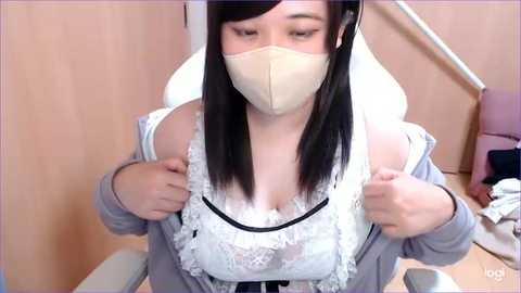 Media: Video of a young Asian woman with fair skin and straight black hair, wearing a white lace bra and beige face mask, adjusting a gray cardigan in a light-wood room with a white chair and pink cushion.