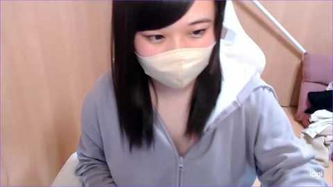 Media: Video of an Asian woman with straight black hair, wearing a light blue hoodie, and a white surgical mask, sitting in a chair with a wooden headrest, in a hospital room with a pink chair and white sheets.