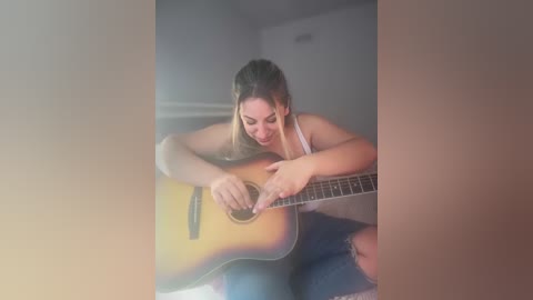 Media: Video of a young woman with long hair, wearing a white tank top and jeans, playing an acoustic guitar in a dimly lit room with soft, warm lighting.