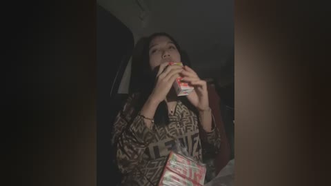 Media: A dimly-lit video of a young woman with long black hair, wearing a patterned top, eating from a fast food bag, surrounded by dark, shadowy interior.