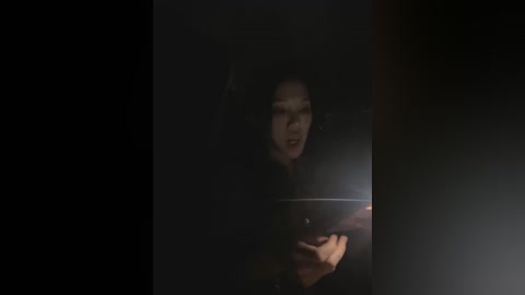 Media: A dimly lit video shows a man with short, dark hair, wearing a dark shirt, holding a smartphone, partially illuminated by the screen's light. The background is dark and indistinct.