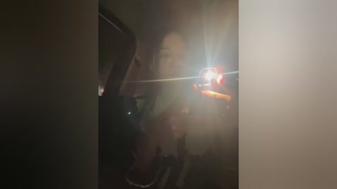 Media: A dimly lit video of a person with long hair and a black top, taking a selfie in a mirror, with a bright flash causing a glare and blurring the reflection.