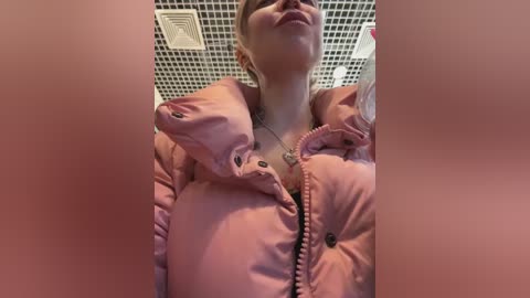 Media: Video of a pregnant woman with blonde hair, wearing a pink puffy jacket, smiling. The background features a tiled ceiling with a ventilation grill.