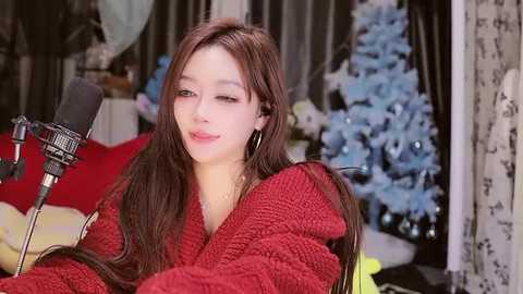 Media: A video of a smiling, light-skinned woman with long brown hair, wearing a red knit sweater and gold hoop earrings, singing into a black microphone. Background features a Christmas tree and a bed with red sheets.