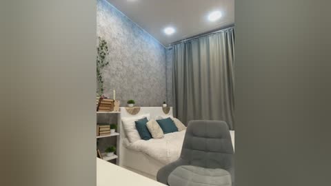 Media: Video of a modern, minimalist bedroom with a white bed adorned with teal pillows, gray curtains, a gray chair, and a potted plant on a white shelf.