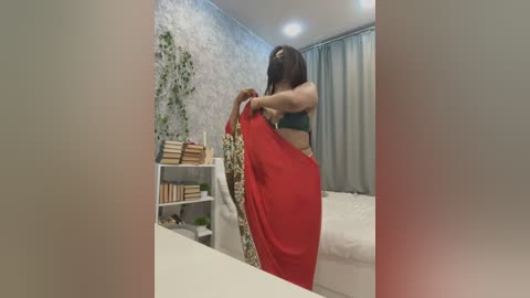 Media: Video of a woman in a green top, lifting a red saree in a modern bedroom with white walls, grey curtains, and a bookshelf.