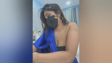 Media: Video of a young woman with medium brown skin, long black hair, wearing a black face mask, a blue robe, and large silver hoop earrings, posing in a modern bedroom.