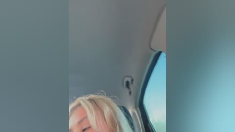 Media: Video of a blonde woman's face, partially cropped, inside a car. She has light skin and is wearing a blue shirt. The background shows the car's interior, with a window reflecting blue sky.