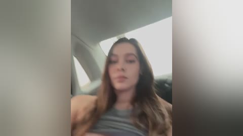 Media: Video of a young woman with long brown hair and light skin, wearing a gray top and beige jacket, sitting in a car with light gray interior, blurred background.