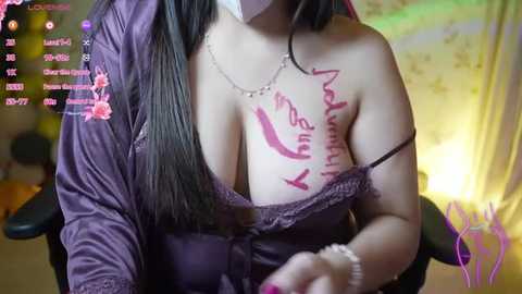 Media: Video of a curvy woman with long black hair, wearing a purple satin robe, revealing a large breast with pink tattoos, sitting in a dimly lit room with a yellow curtain and video game controls.