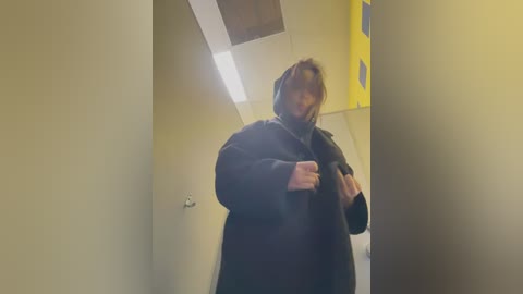 Media: A video shows a woman in a black coat and hood walking down a dimly lit hallway with yellow walls and a bright overhead light.