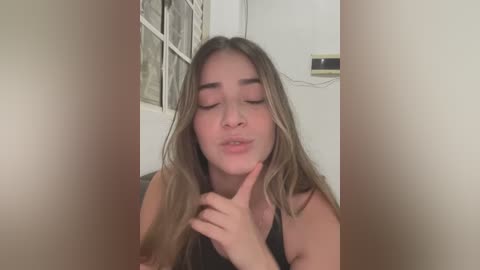 Media: Video of a young woman with long, wavy, light brown hair, wearing a black sleeveless top, making a heart shape with her fingers while closing her eyes. The background features a white wall and a window with frosted glass.