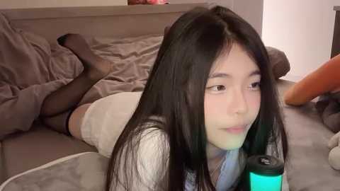 Media: Video of an Asian woman with long black hair, lying on a bed in a casual outfit, holding a glowing green drink, in a cozy bedroom.