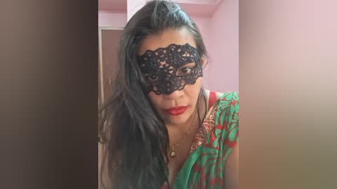 Media: Video of a woman with long black hair, wearing a black lace masquerade mask and a green sari with red floral patterns, in a pink room.