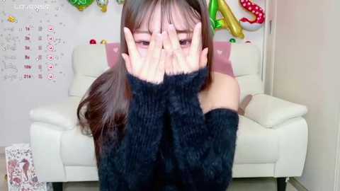 Media: Video of an Asian woman with long dark hair, wearing a black off-shoulder sweater, covering her face with her hands, sitting on a white couch in a brightly lit room with colorful decor.