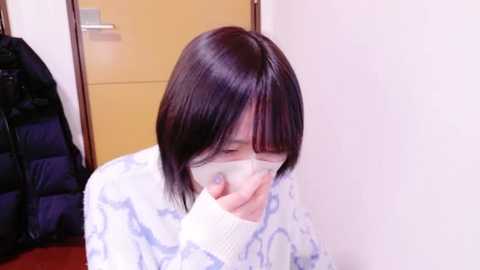 Media: Video of an Asian woman with short black hair and glasses, wearing a white sweater with blue patterns, holding her face, standing indoors near a closed door and a black jacket.