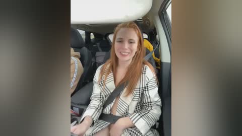 Media: Video of a smiling, fair-skinned woman with long red hair, wearing a plaid dress, sitting in a car seatbelt, surrounded by the car's interior.