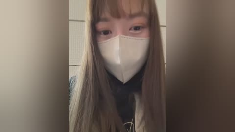 Media: A video of a young woman with long, straight, light brown hair, wearing a white surgical mask and a black top, captured from a low angle, with blurred background of beige walls and a door.