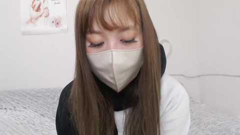 Media: A video of an Asian woman with long, straight brown hair, wearing a beige face mask and a black scarf over a white shirt. She's indoors with a light gray bedspread and a poster on the wall.