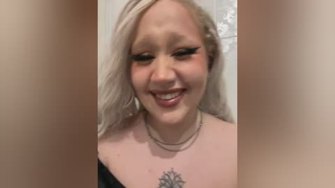 Media: Video of a young, smiling woman with platinum blonde hair, wearing black lingerie, and a delicate necklace. She has a small, intricate tattoo on her collarbone. The background is blurred, focusing attention on her.