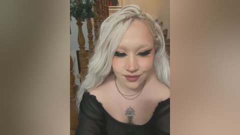 Media: Video of a young woman with long, platinum blonde hair, wearing black clothing, and a necklace with a large pendant. She has a small tattoo on her chest. Background features wooden furniture and a plant.