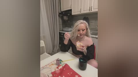 Media: Video of a blonde woman in a kitchen, eating cake, wearing a black top, pink pants, sitting at a table with a red cloth, green cup, and utensils.