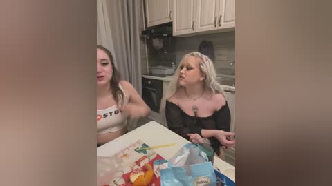 Media: A video of two women in a kitchen, one in a white Hooters crop top, the other in a black off-shoulder top, holding groceries, beige cabinets, and a white countertop.