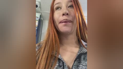 Media: Video of a Caucasian woman with long, vibrant orange dreadlocks, wearing a gray and white striped shirt, looking into the camera with a slight smile. Background shows a blurred hospital setting.