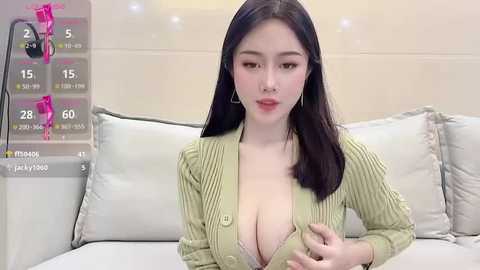 Media: Video of an East Asian woman with long black hair, fair skin, and large breasts, wearing a low-cut, ribbed, green cardigan, sitting on a white bed, surrounded by white pillows, in a modern bedroom.