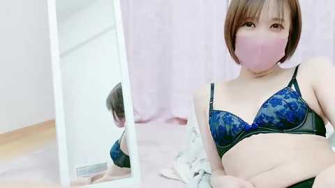 Media: Video of an Asian woman with short brown hair and a pink face mask, wearing a blue lace bra, sitting on a bed with a mirror reflecting her partially.