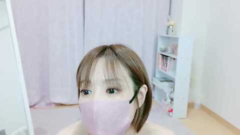 Media: A video of a young Asian woman with straight, shoulder-length brown hair, wearing a pink surgical mask and black headphones, sitting indoors in a minimalistic room with light wooden floors and pastel curtains.