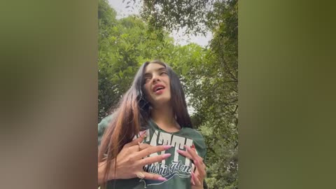 Media: Video of a young woman with long brown hair, wearing a green t-shirt, smiling and making heart shapes with her hands, surrounded by lush green foliage.