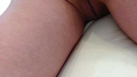 Media: Video of a close-up of a light-skinned person's lower torso and thigh, with visible skin texture and a black thong partially visible. The background is a white surface, likely a bed or couch.