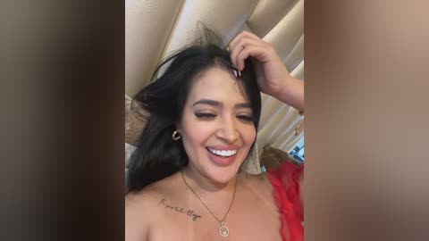 Media: Video of a smiling Latina woman with dark hair, wearing a red dress, and a necklace, adjusting her hair. Background shows a ceiling and window blinds.