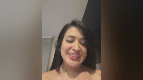 Media: A close-up video of a young, smiling woman with long black hair, wearing a gold necklace. She has a light skin tone and is indoors, possibly in a bedroom, with a white ceiling and a dark object partially visible in the background.