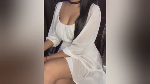 Media: Video of a woman with long black hair, wearing a sheer white dress with a black choker, sitting on a chair in a dimly lit room.