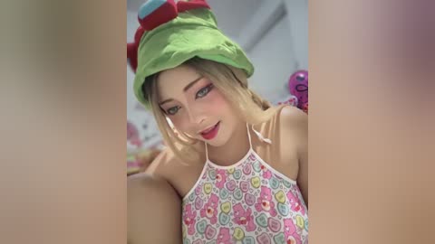 Media: Video of a smiling Asian woman with fair skin, wearing a green bucket hat and a colorful floral apron, set in a bright, indoor room with a pink toy in the background.