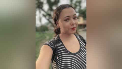 Media: Video of a young woman with light brown skin, wearing a black and white striped top, taking a selfie outdoors. Background features blurred greenery and trees.