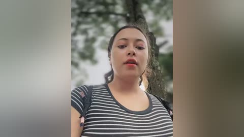 Media: Video of a young woman with light brown skin and curly dark hair, wearing a gray and white striped shirt, standing outdoors in a forest setting with blurred trees in the background.