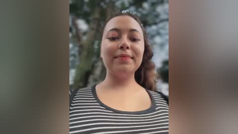 Media: Video of a young woman with light brown skin, straight brown hair, wearing a black and white striped shirt, standing outdoors with blurred green foliage in the background.