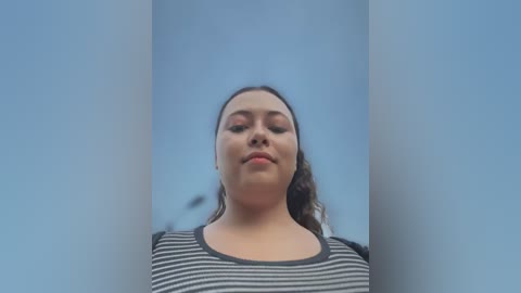 Media: Video of a young woman with medium skin tone and curly dark hair, wearing a black-and-white striped shirt, standing in front of two vertical white columns, creating a minimalist, serene background.