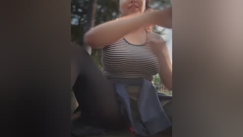 Media: Video of a woman with fair skin, wearing a striped tank top and denim shorts, sitting in a car, surrounded by blurred trees.