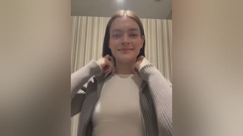 Media: Video of a young woman with fair skin, straight brown hair, and a light gray sweater. She stands in front of beige curtains, smiling slightly, holding her ears with both hands.