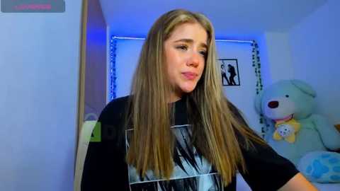 Media: Video of a young woman with long, straight, light brown hair, wearing a black graphic T-shirt. She's in a cozy bedroom with a teddy bear, a blue wall, and a framed picture.