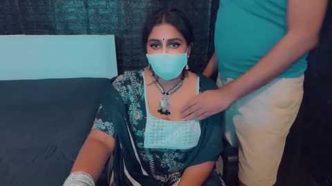 Media: Video of a South Asian woman in a teal sari with a white patterned blouse, wearing a blue surgical mask and a red bindi, being touched by a man in a turquoise shirt and beige pants, in a dimly lit room.