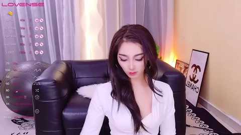 Media: A video of a young East Asian woman with long black hair, wearing a white blouse, sitting on a black leather couch in a modern living room with a TV screen showing \"Love TV\" in the background.