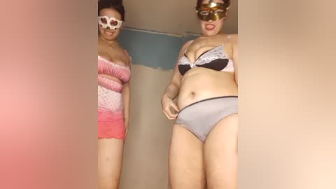 Media: Video of two women in lingerie and masks in a dimly-lit room, one in pink and the other in grey.