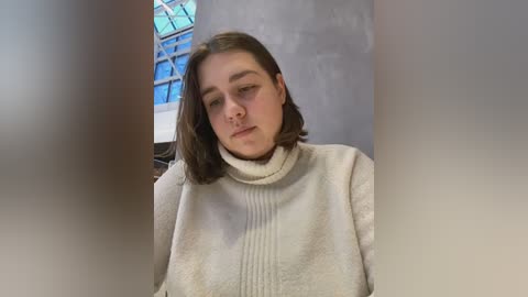 Media: Video of a young woman with shoulder-length brown hair, wearing a beige turtleneck sweater, sitting indoors with a blue glass ceiling and concrete wall in the background.